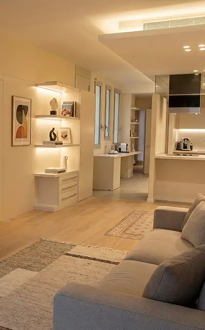 Renovations for Expats Barcelona showing a warm modern open-plan living room with built-in shelves, sofa, wood flooring and integrated kitchen - by Revive Quality